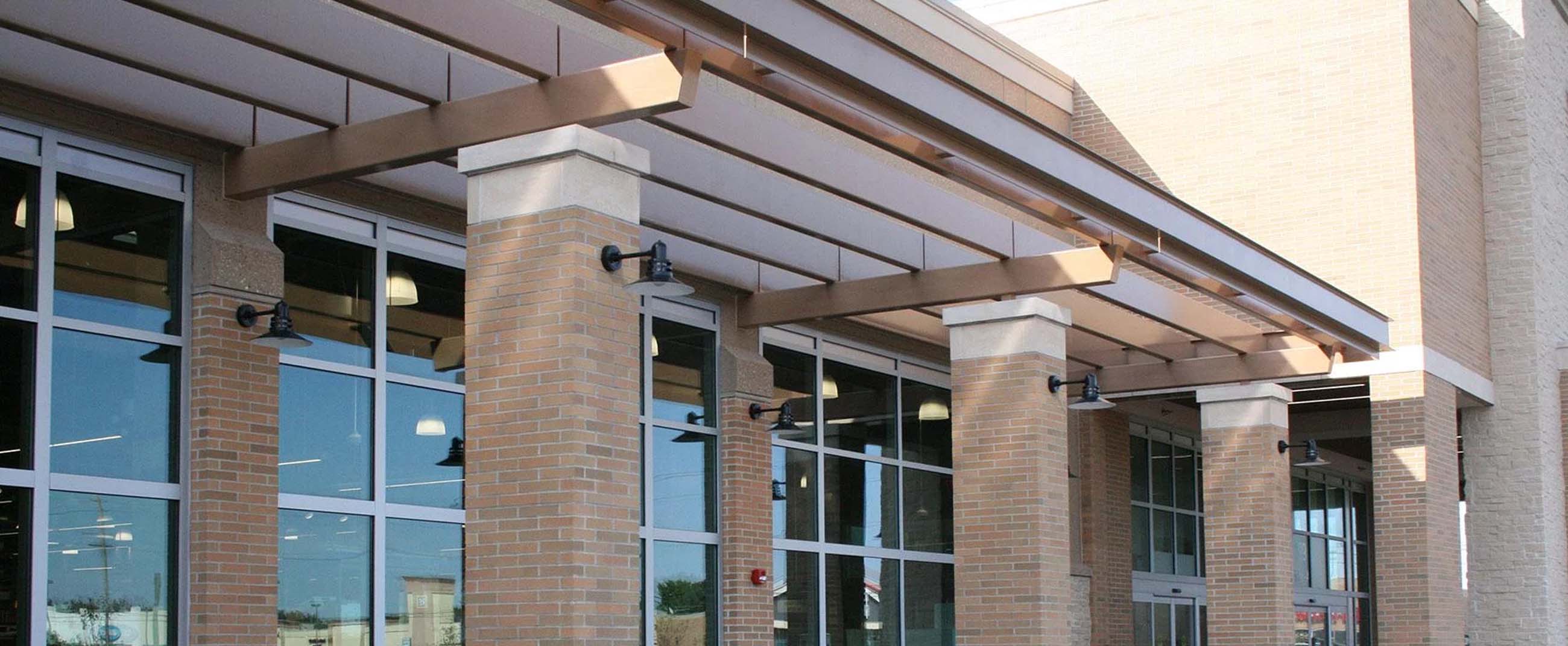 the facade of a modern commercial building featuring a brick structure, large windows, and columns supporting a metal canopy. The design incorporates landscaped areas and outdoor seating, creating a pleasant and functional space for customers and visitors.
