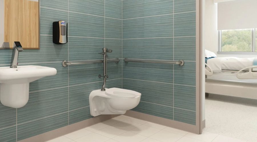 Baño accesible con lavabo mural, dispensador de jabón, barras de apoyo y sanitario suspendido; se aprecia una habitación de hospital al fondo.