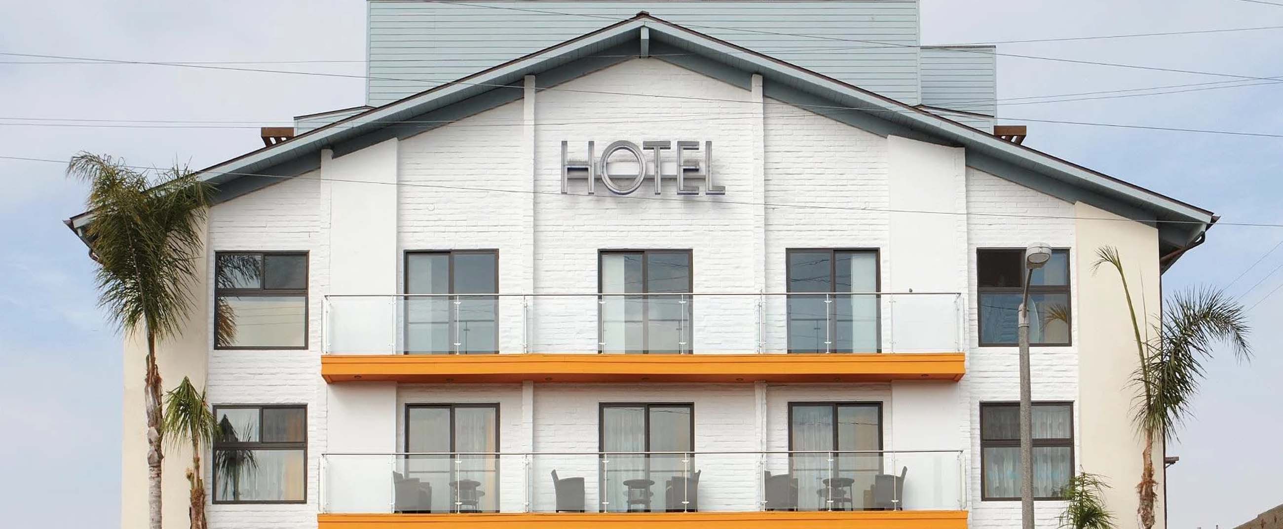 Hotel Erwin, an iconic property located in Venice Beach, Los Angeles. The building features a modern, laid-back design with white walls, glass balconies, and bright orange accents that capture the lively beach atmosphere. Palm trees frame the exterior, enhancing the coastal California vibe, while the bold metallic “HOTEL” sign underscores its minimalist and contemporary style.