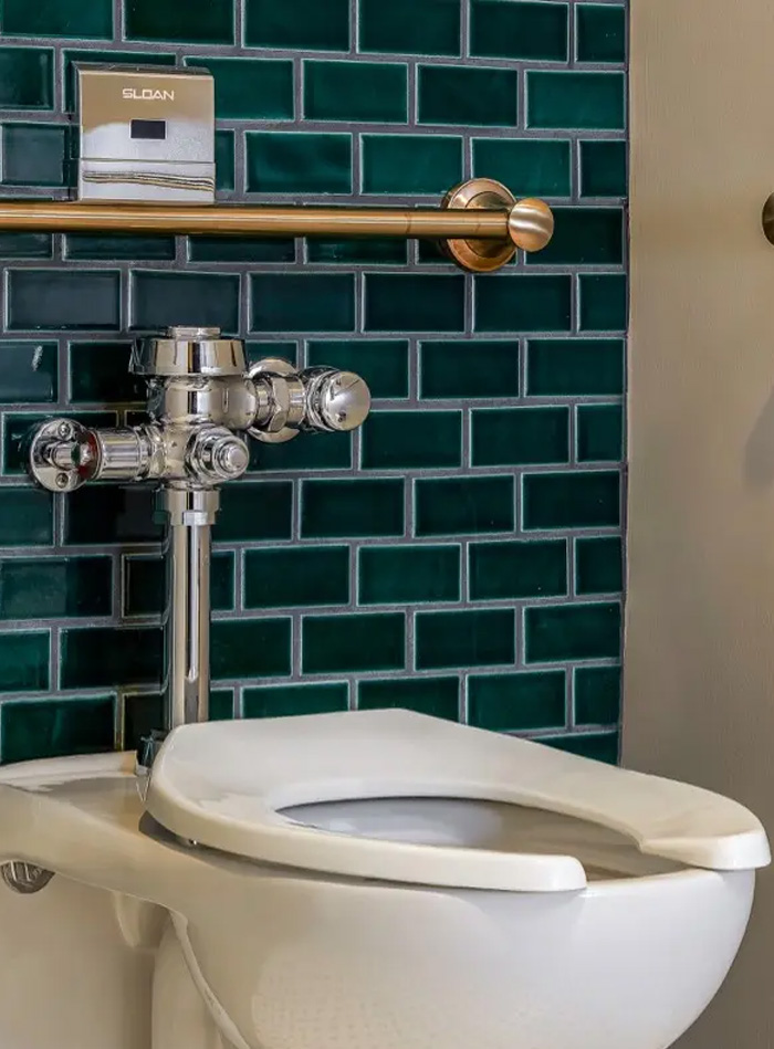 toilet fitted with a Sloan chrome flushometer, set against a wall of green subway tiles with light grout lines that add texture and sophistication. Above it, a gold grab bar enhances accessibility while maintaining a refined appearance. Located at the Hotel Zachary, this design perfectly blends contemporary elegance with classic comfort, consistent with the hotel’s signature style.