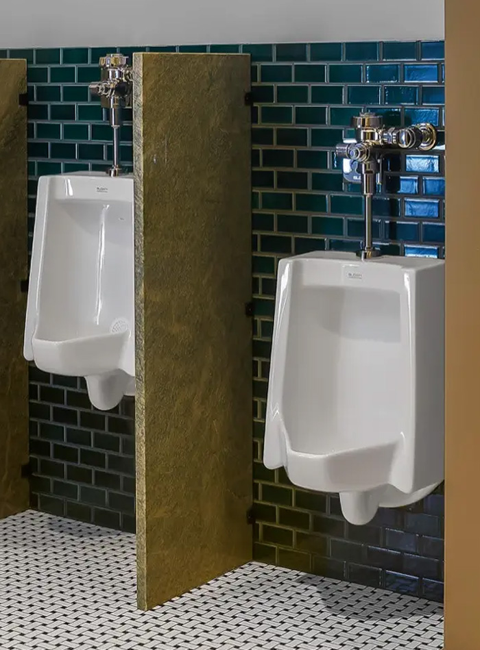 a set of urinals with Sloan chrome flushometers, installed in a stylish and modern restroom. The backdrop features emerald-green subway tiles, providing depth and contrast, while the green marble dividers add a refined touch. The black-and-white mosaic floor pattern complements the timeless yet contemporary aesthetic of the Hotel Zachary, balancing functionality and sophisticated design.