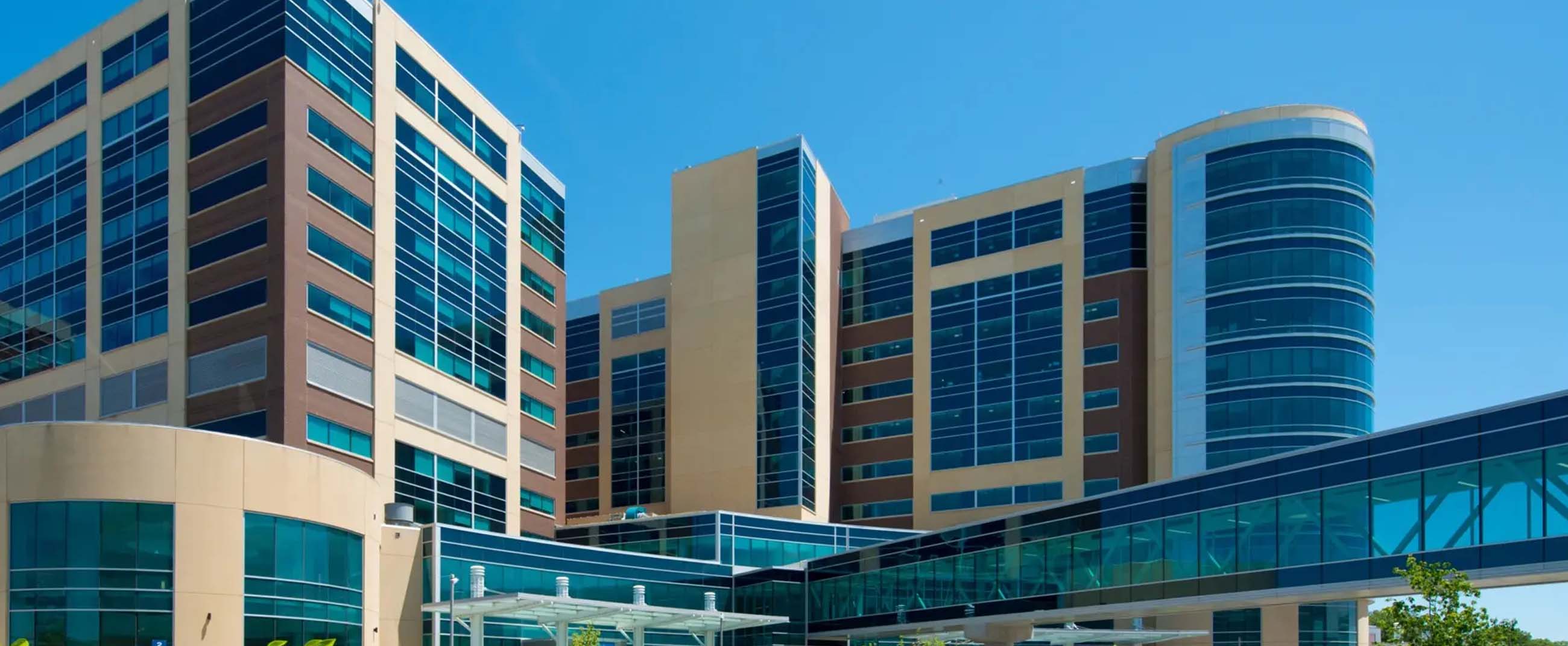 a modern hospital complex with large glass façades and contemporary architectural design. The building features beige and brown tones with blue-tinted windows that reflect the sky, conveying a sense of cleanliness, innovation, and efficiency. In the foreground, landscaped greenery and a driveway lead to the main entrance with a glass canopy.