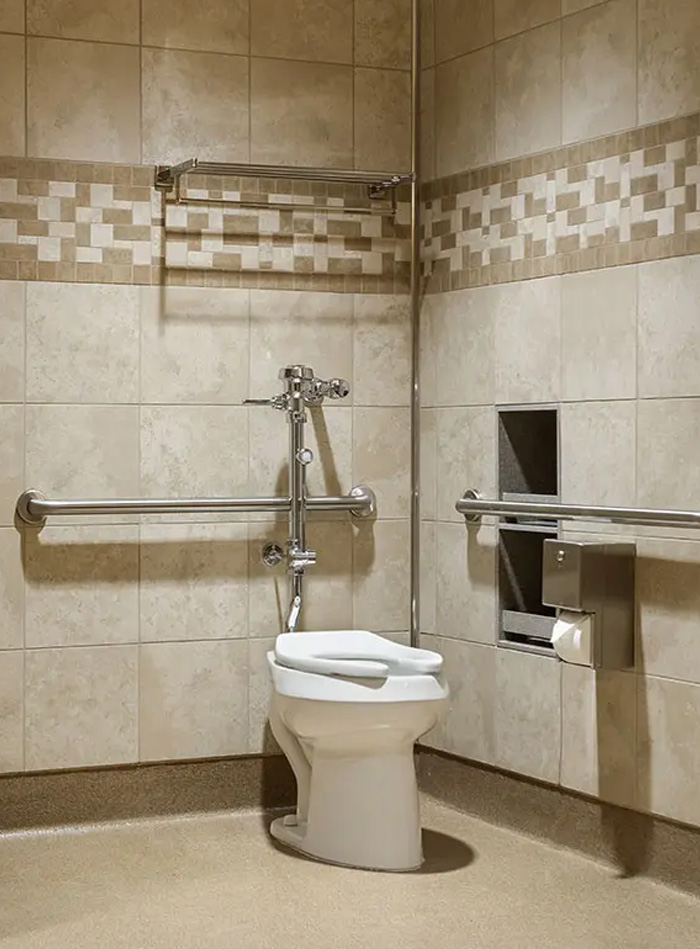 a white ceramic toilet with an open-front seat, installed in a restroom stall lined with beige tiles. The chrome flushometer adds a modern and functional touch, while the neutral finishes and lighting create a clean and orderly atmosphere.