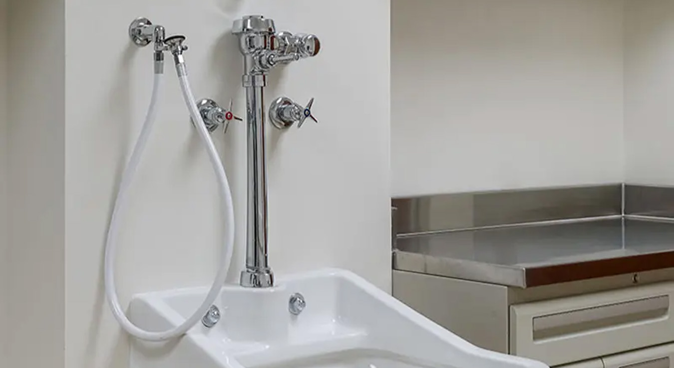 a chrome flushometer, hot and cold water control valves, and a flexible rinse hose. The clean environment, featuring stainless steel surfaces and built-in cabinets, highlights a functional and hygienic space ideal for hospital or laboratory settings.