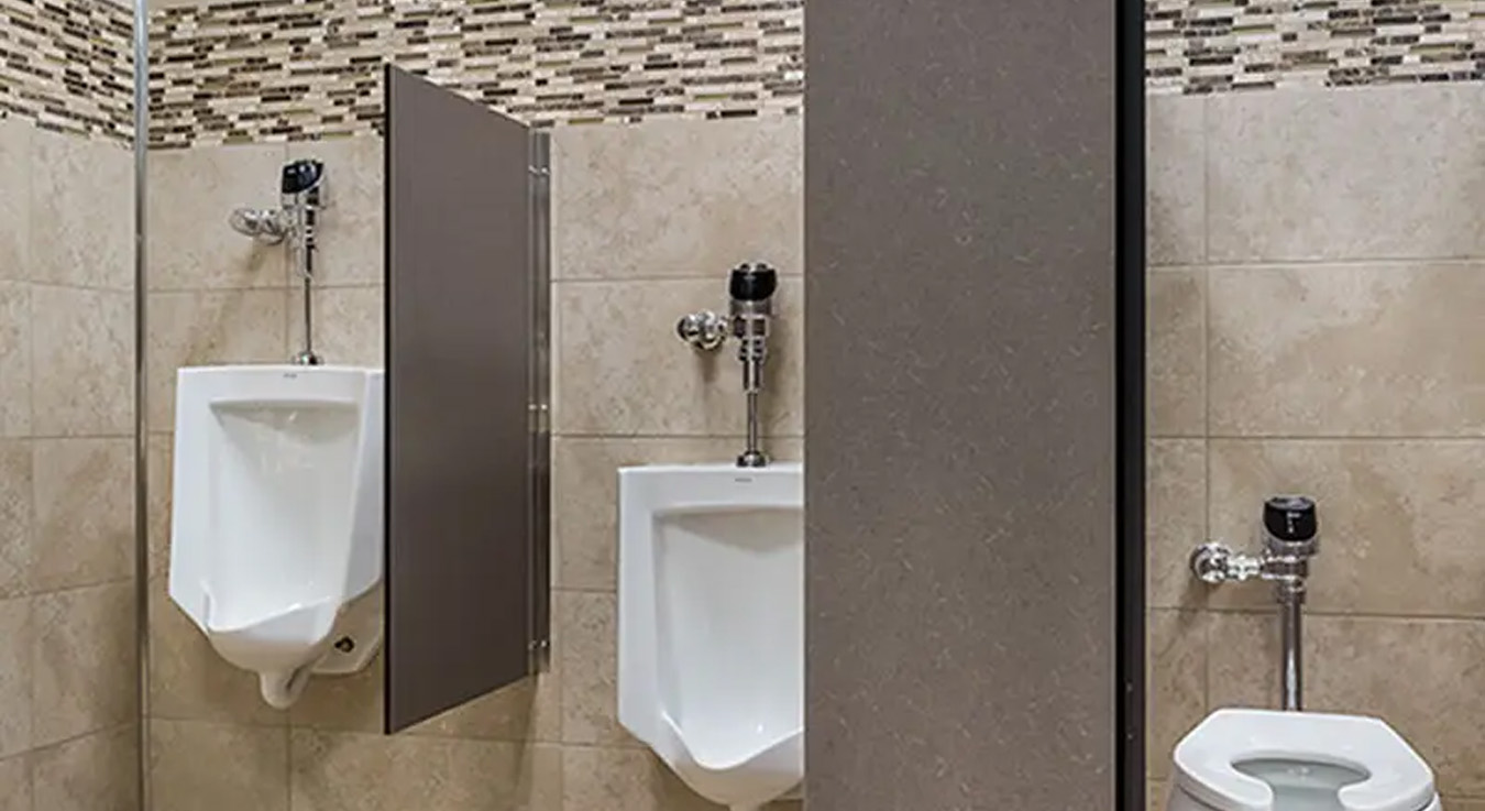 a modern public restroom with beige finishes and decorative mosaic details on the walls. Two urinals and one toilet are visible, all equipped with chrome automatic flushometers. The clean design, balanced lighting, and discreet partitions reflect a functional, hygienic, and well-maintained environment.