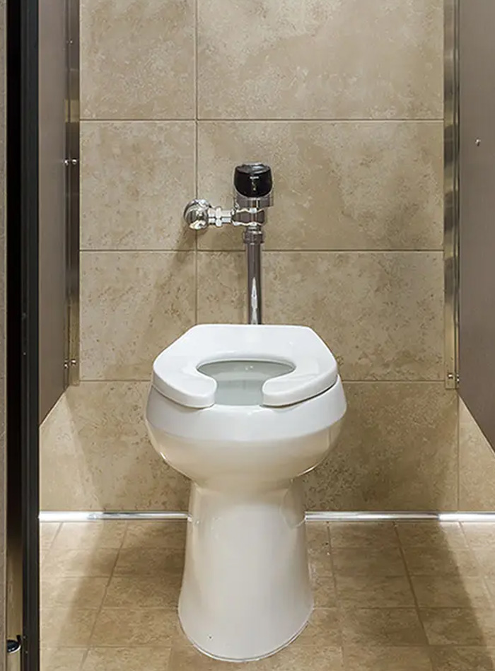 a white ceramic toilet with an open-front seat, installed in a restroom stall lined with beige tiles. The chrome flushometer adds a modern and functional touch, while the neutral finishes and lighting create a clean and orderly atmosphere.
