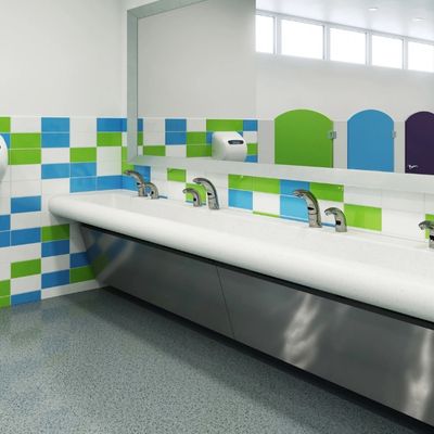 Lavabo largo de superficie sólida con grifos automáticos Sloan, instalado frente a una pared con azulejos verdes, azules y blancos en un baño escolar moderno.