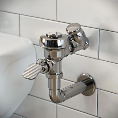 Sloan manual urinal flushometer with pedal in polished chrome finish, installed against a white subway tile wall.