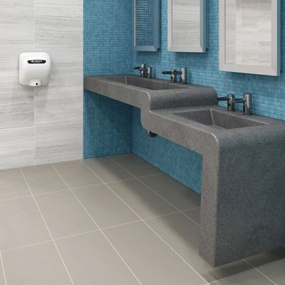 Lavabo doble de superficie sólida gris con grifos automáticos Sloan, frente a una pared de mosaico azul con un secador de manos a un costado.