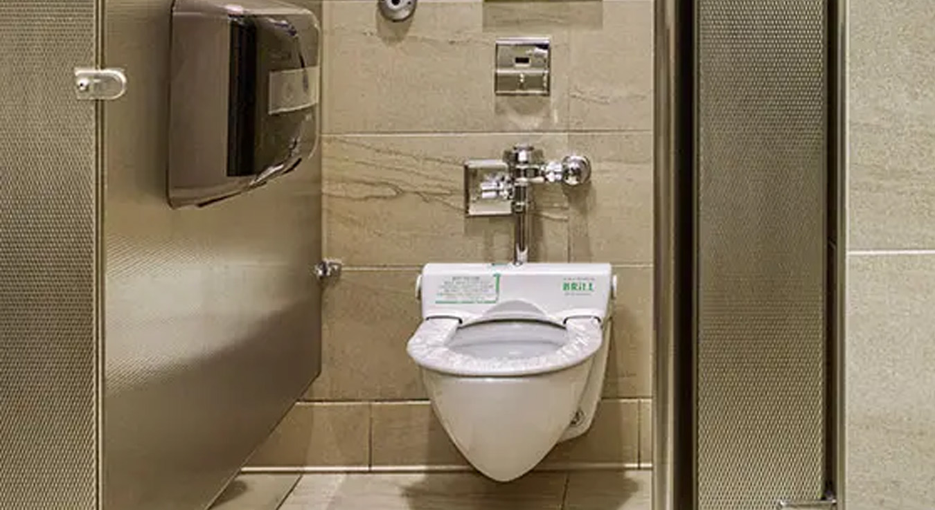 a modern public restroom with beige and metallic finishes. In the foreground, there is a stall with a wall-mounted toilet featuring a chrome flush valve and an automatic hygienic seat cover. The stall partitions are metallic with a textured surface, and the floor is tiled with ceramic panels.