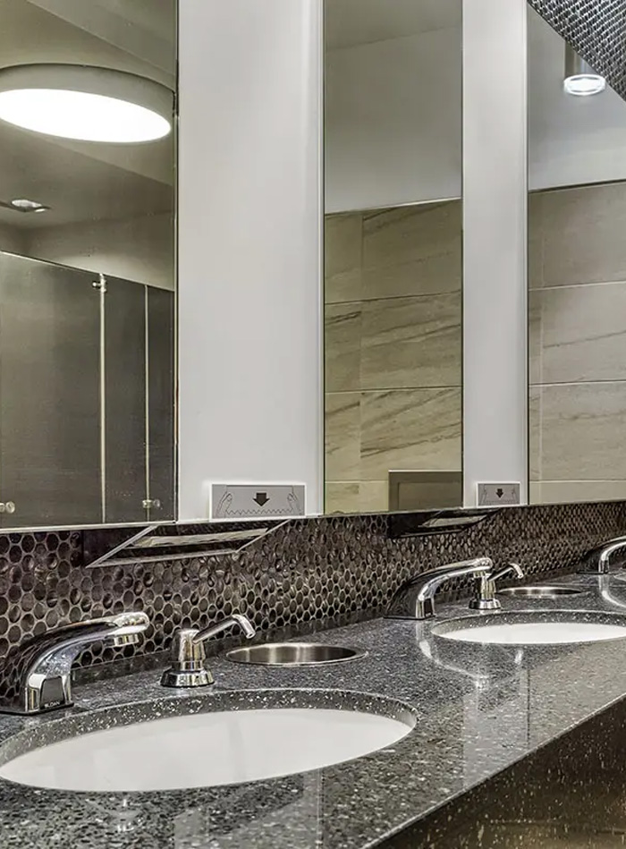 a modern public restroom with beige and metallic finishes. In the foreground, there is a stall with a wall-mounted toilet featuring a chrome flush valve and an automatic hygienic seat cover. The stall partitions are metallic with a textured surface, and the floor is tiled with ceramic panels.