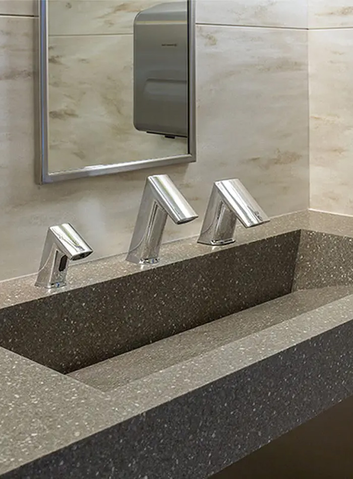 a modern solid-surface sink in dark gray with a speckled texture. It features three chrome automatic faucets with a sleek angular design, positioned beneath a rectangular mirror framed in metal. The walls are covered in light beige polished tiles, and a stainless steel towel dispenser can be seen in the background.