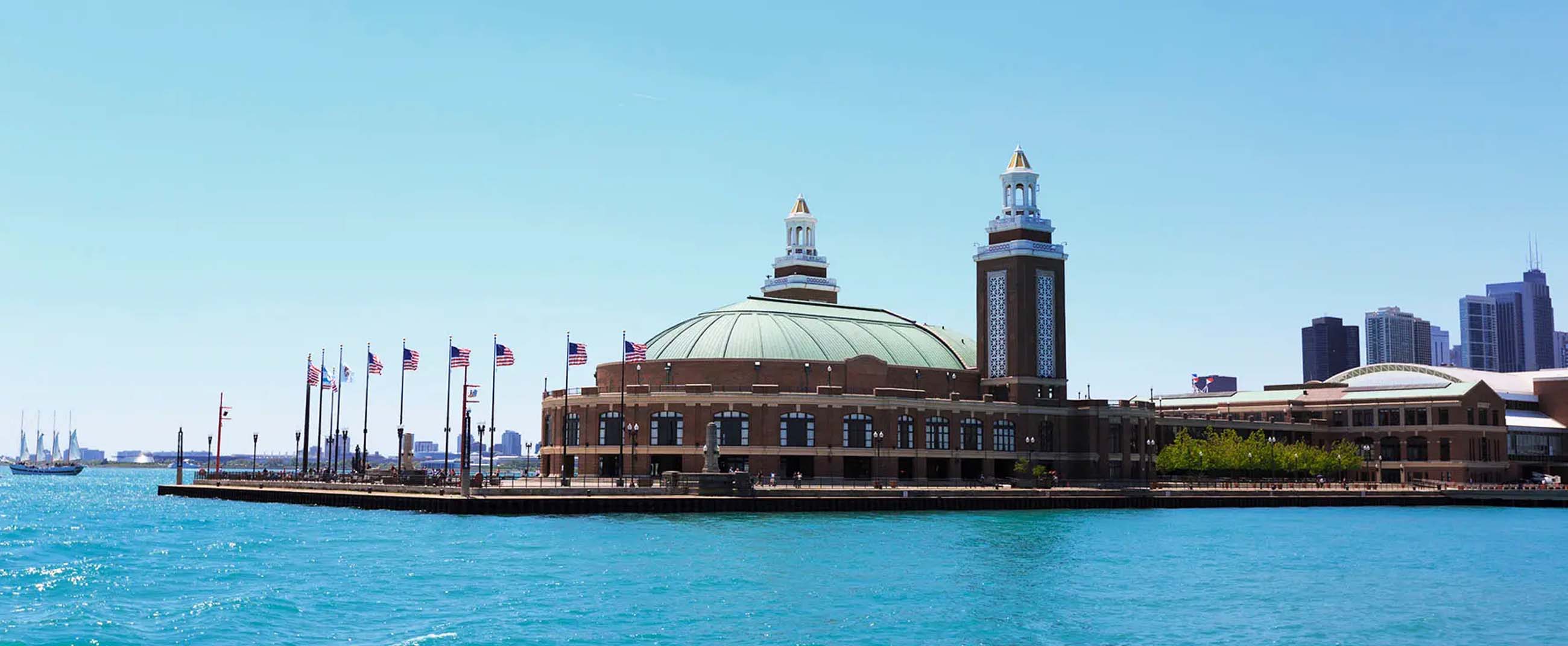 Navy Pier in Chicago, Illinois — one of the city’s most iconic tourist landmarks. The main building, with its green dome and twin brick towers, extends over Lake Michigan, surrounded by a pier lined with multiple U.S. flags waving in the breeze. The turquoise water and clear sky highlight the architectural elegance of the venue, which blends history and modernity as it hosts events, restaurants, and cultural attractions.