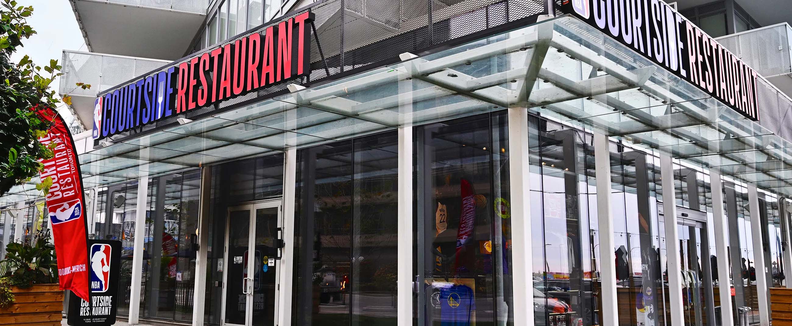 the exterior of the NBA Courtside Restaurant, a modern, NBA-themed dining venue. The building features glass and metal architecture with clean lines, highlighted by a bold sign displaying the league’s official logo and the restaurant name. The overall design conveys an urban, contemporary feel, perfect for basketball fans seeking a sports-inspired dining experience.