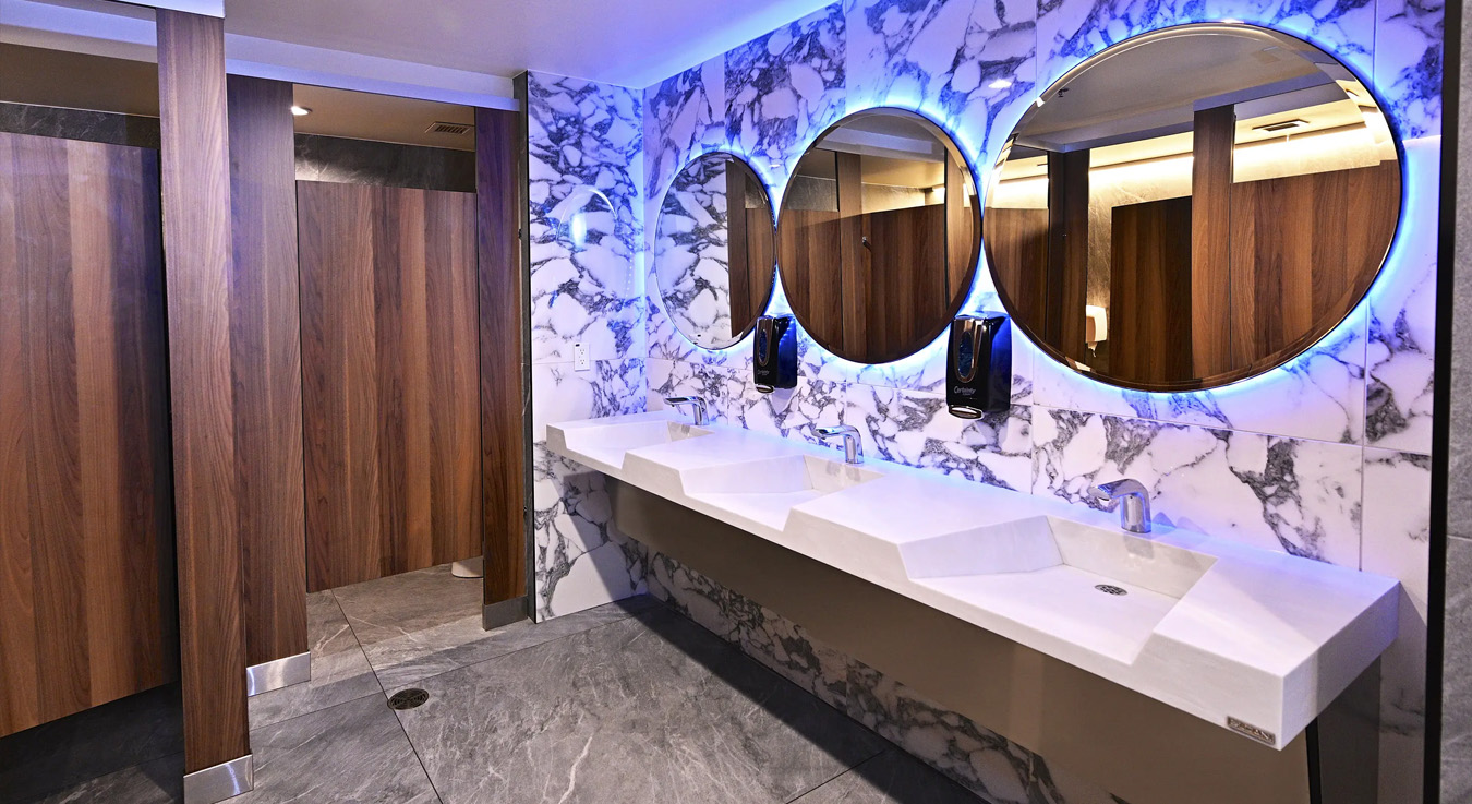 stylish restroom at the NBA Courtside Restaurant, blending contemporary luxury with hygienic functionality. The solid-surface sink features Sloan touchless faucets, automatic soap dispensers, and round mirrors with blue backlighting, creating a sleek, high-tech atmosphere. The combination of veined white marble and warm wood finishes enhances the refined aesthetic, perfectly matching the restaurant’s premium brand identity.
