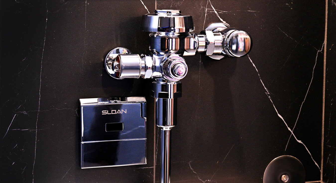 a Sloan flushometer mounted on a black marble wall with white veining, embodying a sleek and contemporary aesthetic. Its polished chrome finish and integrated infrared sensor emphasize touch-free technology, perfectly suited for maintaining high hygiene and efficiency standards in upscale environments such as the NBA Courtside Restaurant.