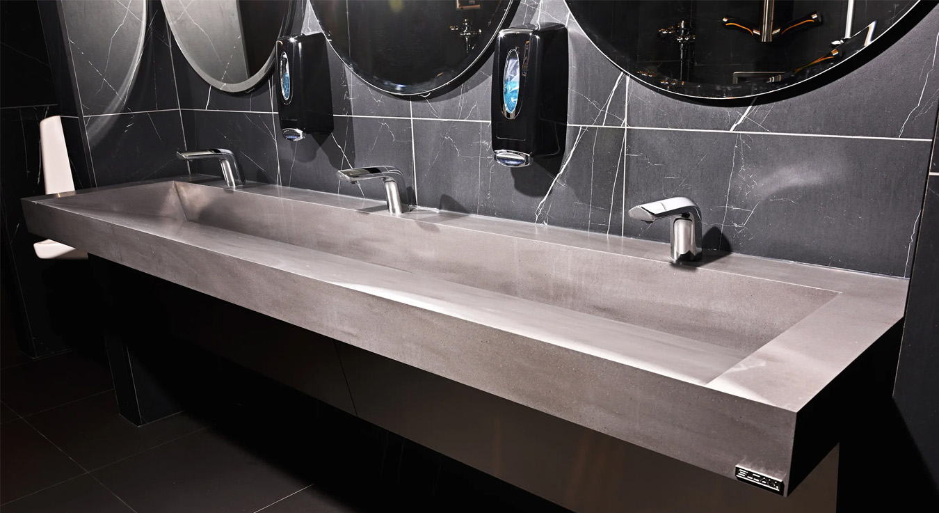a stainless steel sink station featuring Sloan touch-free faucets, set against a sleek black marble backdrop with round, backlit mirrors. The design merges modern luxury and hygienic efficiency, perfectly fitting the atmosphere of the NBA Courtside Restaurant