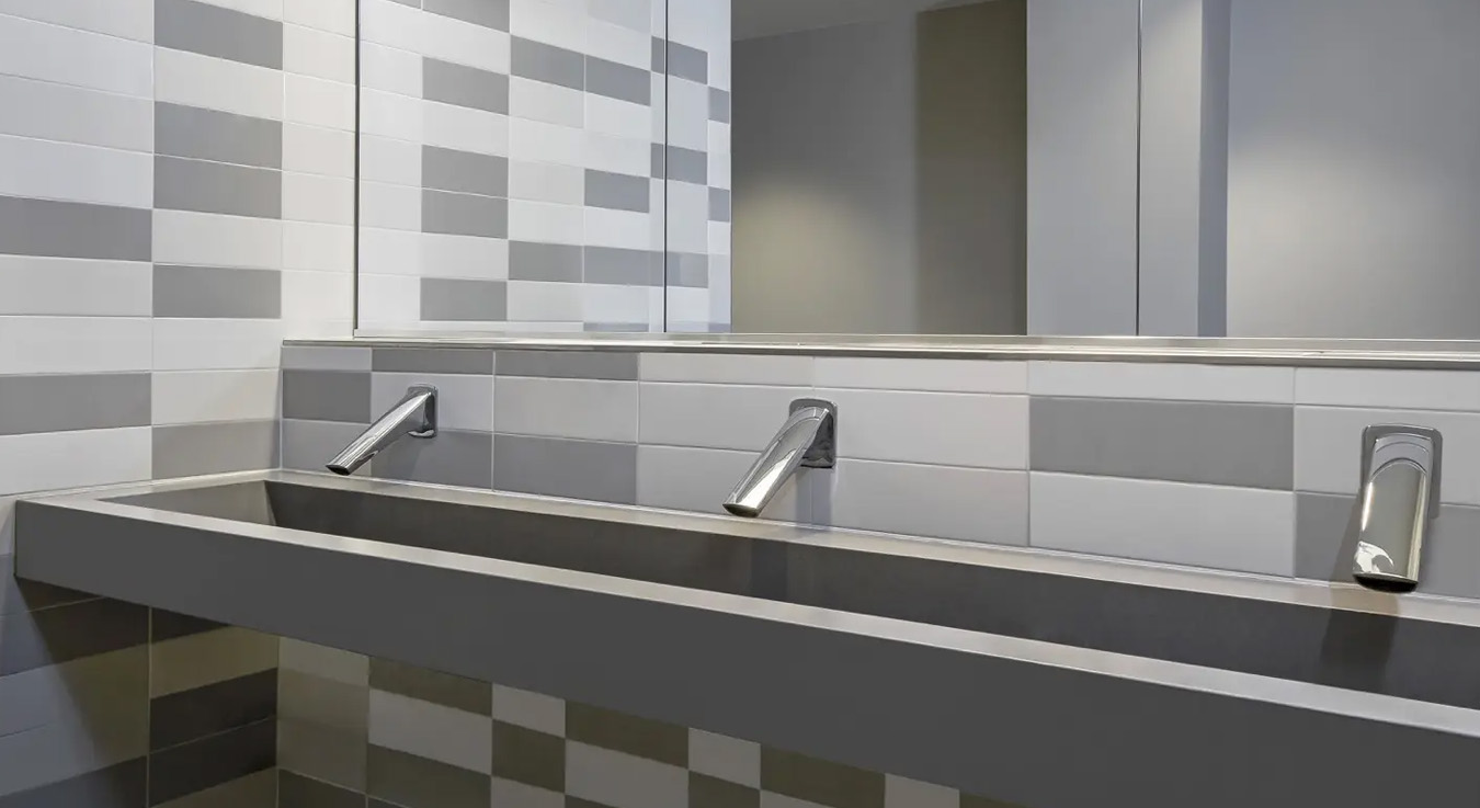 stainless steel trough-style sink with wall-mounted automatic faucets, designed for multiple users simultaneously. The background features gray and white tiles arranged in a geometric pattern, reinforcing the restroom’s modern aesthetic.