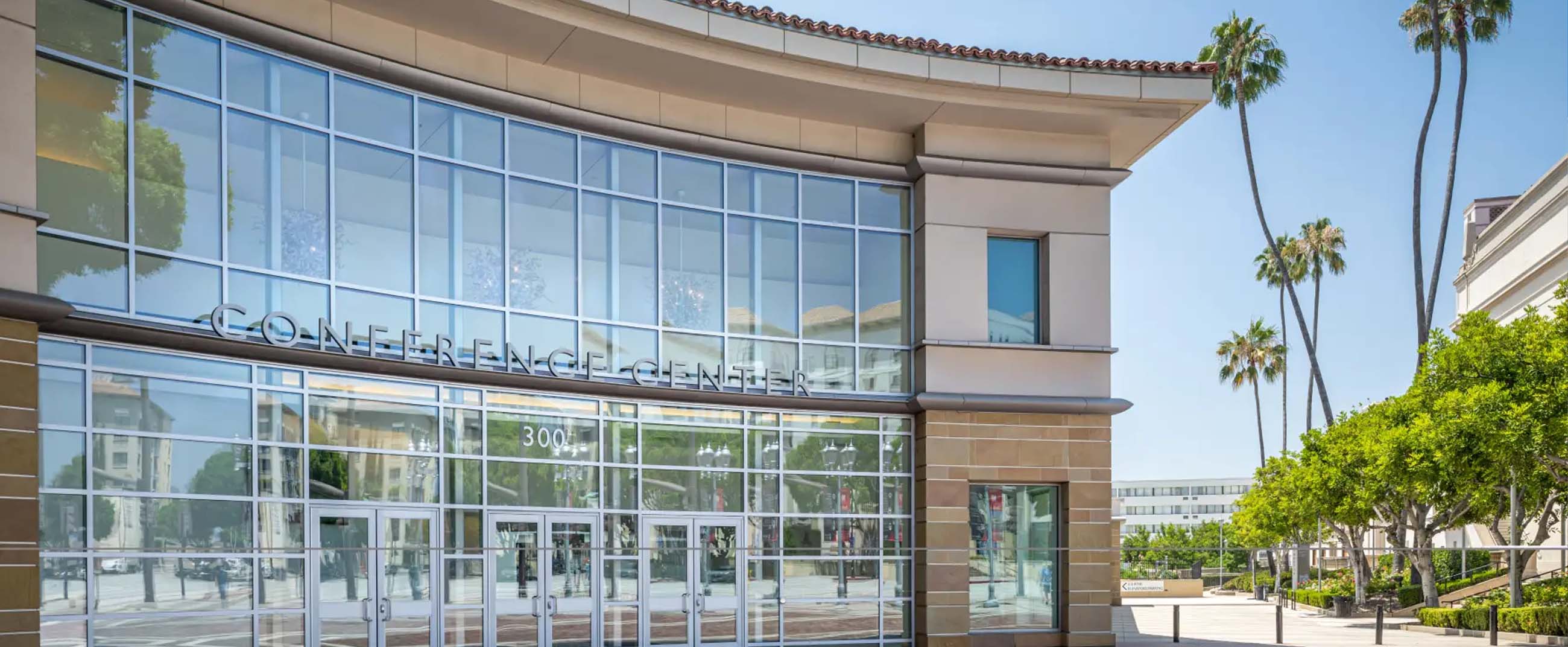 Pasadena Convention Center in California, featuring a modern design with large glass windows and a metal framework that allows abundant natural light. The building’s curved lines, light stone panels, and neutral tones blend seamlessly with the urban surroundings. In front, a brick-paved plaza and tall palm trees frame the main entrance, conveying a bright, orderly atmosphere characteristic of contemporary Californian architecture.