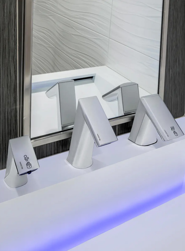 close-up of a modern sink featuring Sloan automatic chrome faucets with angular lines. The faucets are mounted on a white surface illuminated by blue LED lighting, emphasizing a sleek and high-tech design. A metal-framed mirror and textured white and gray walls complete the contemporary aesthetic, reflecting a clean, functional, and sophisticated restroom environment.