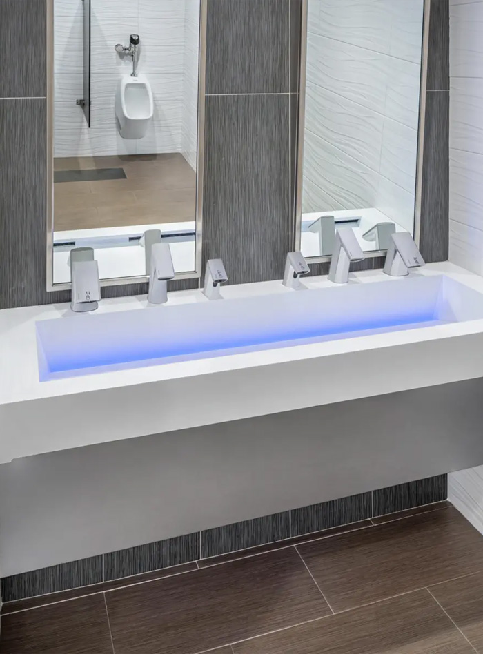 modern white solid-surface sink featuring integrated blue LED lighting that accentuates its contemporary design. Four chrome automatic faucets are aligned in front of vertical mirrors that visually divide the space. The walls combine white and gray textured tiles, while the wood-toned ceramic floor adds warmth. A urinal visible in the reflection indicates a public restroom with a sleek, functional, and technology-driven design.