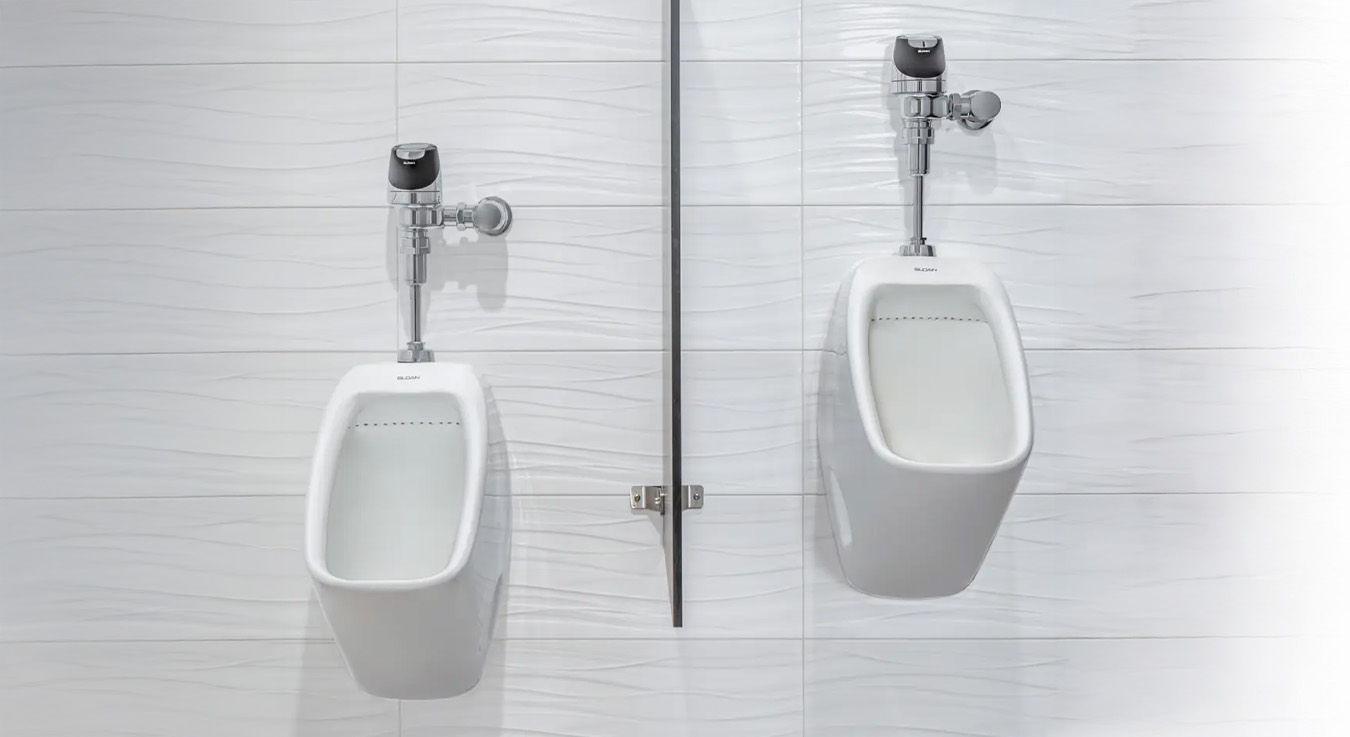 two white Sloan porcelain urinals mounted on a wall clad with white textured tiles featuring a subtle wave pattern. Each unit includes a chrome automatic flush valve and is divided by a slim metal privacy panel that maintains visual symmetry. The bright lighting and modern materials convey a clean, functional, and contemporary restroom design.