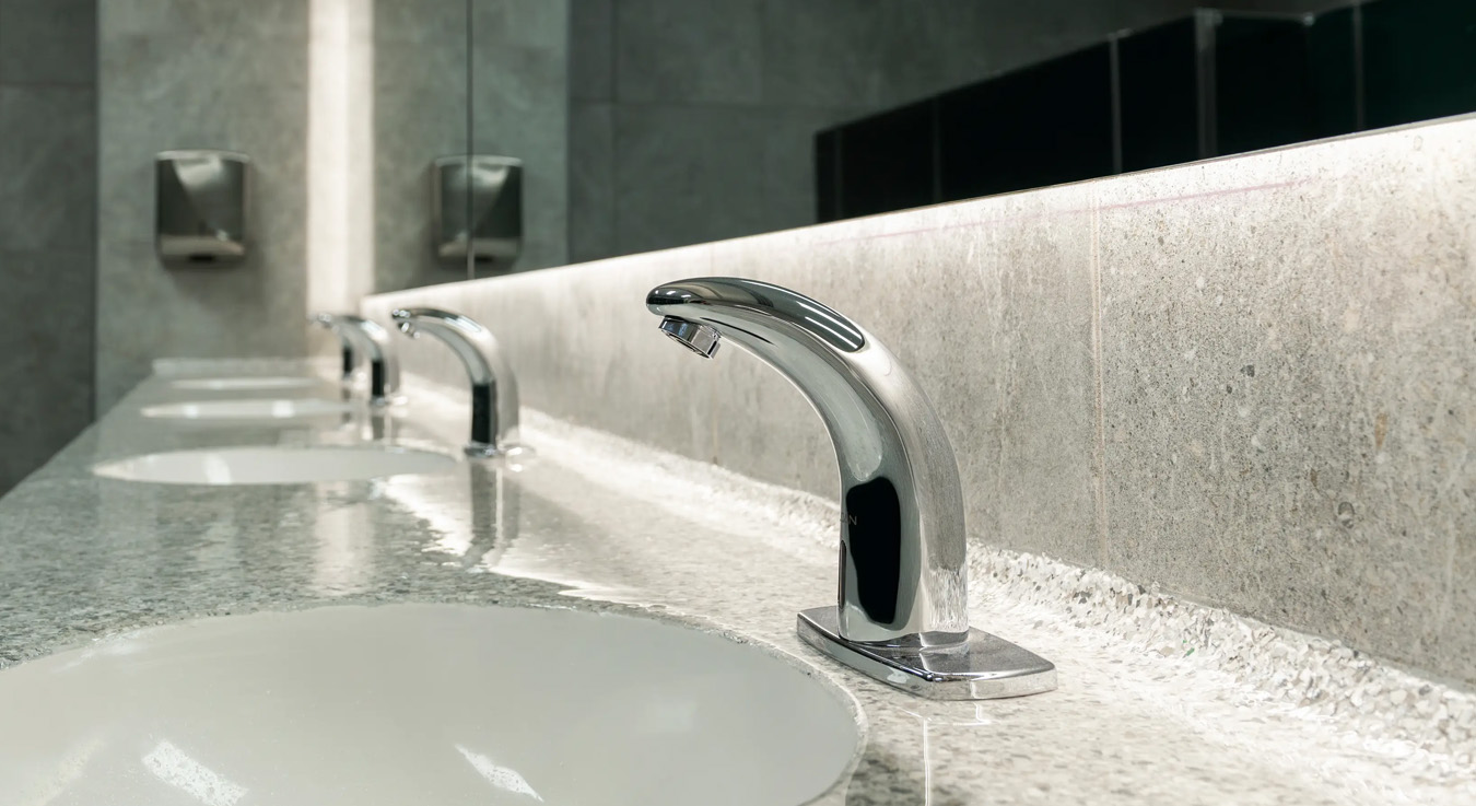 a row of sinks with chrome-finished automatic faucets set on a gray granite countertop. The elegant and minimalist design, complemented by soft lighting, reflects a modern and functional space focused on hygiene and efficiency.