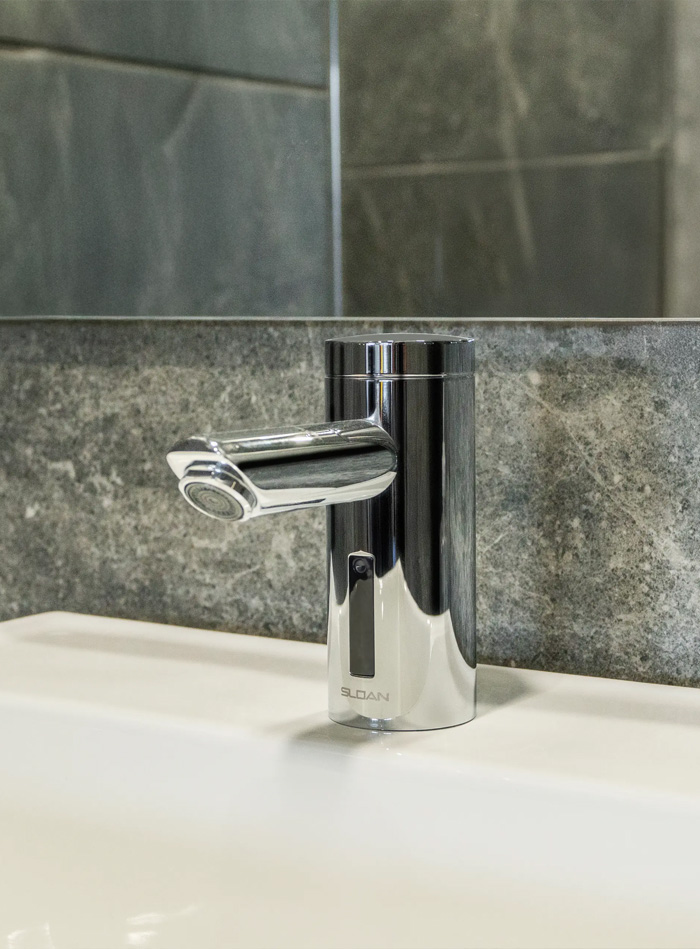 an elegant chrome-finished automatic faucet installed over a white sink. The faucet’s modern and minimalist design, paired with the gray tiled background, creates a clean, sophisticated, and functional atmosphere.