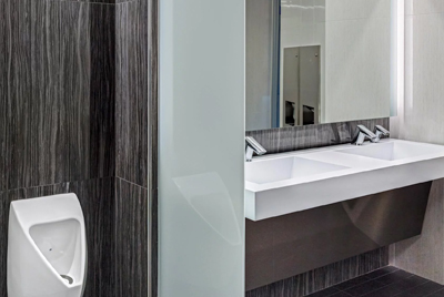 a modern commercial restroom featuring a minimalist design and high-quality materials. The double sink with Sloan® automatic faucets and metallic finishes blends functionality with elegance, while the white porcelain urinal and dark marble panels create a refined visual contrast.