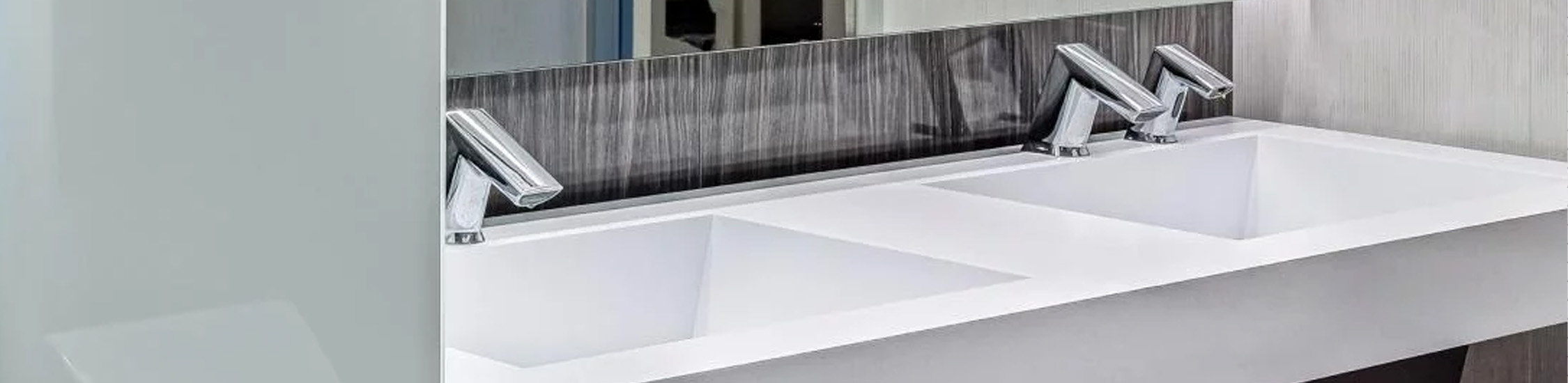 a modern commercial restroom featuring a minimalist design and high-quality materials. The double sink with Sloan® automatic faucets and metallic finishes blends functionality with elegance, while the white porcelain urinal and dark marble panels create a refined visual contrast.