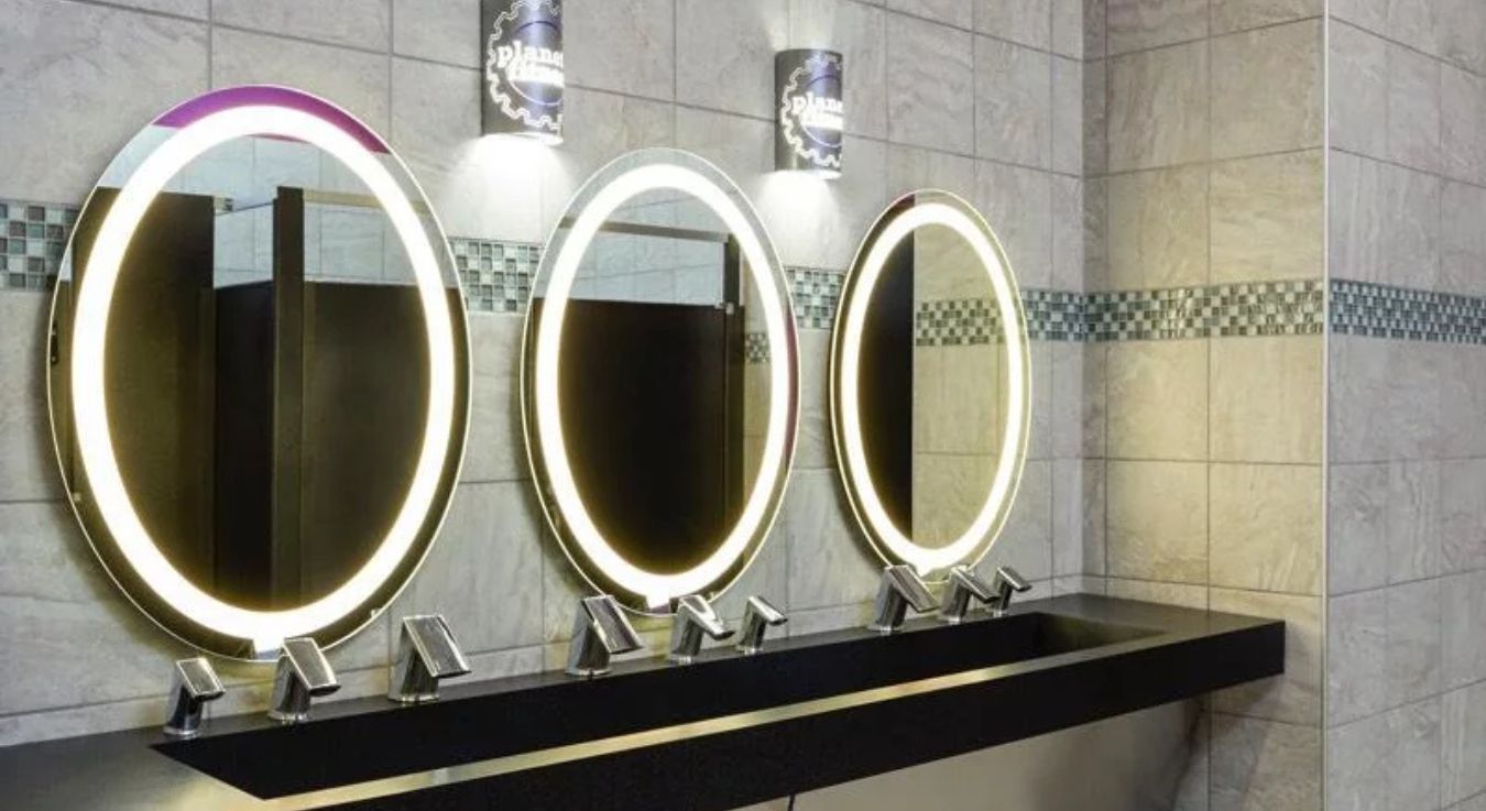 Modern sinks with illuminated circular mirrors and Sloan automatic faucets inside a Planet Fitness restroom designed for comfort, style, and efficiency.