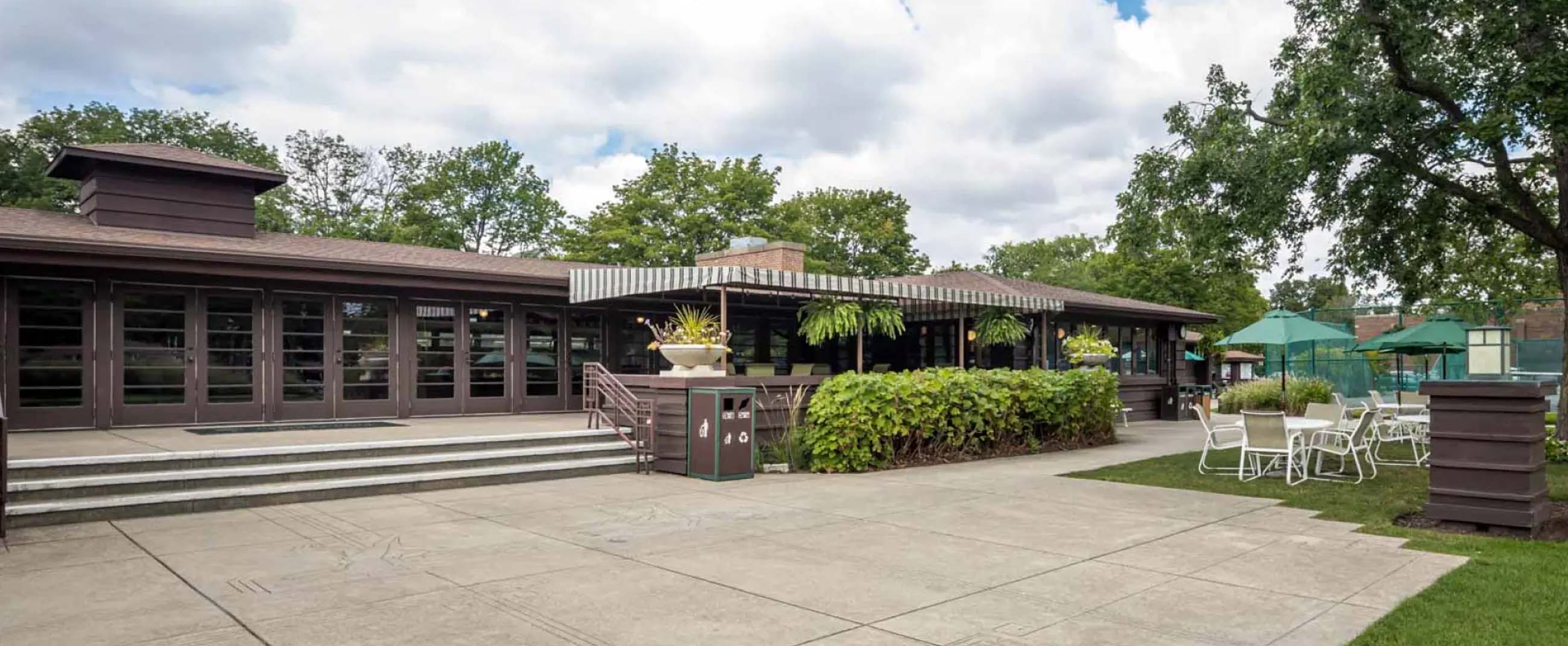 River Forest Tennis Club, a classic American-style building with a low, horizontal structure, large windows, and dark wood finishes. The surroundings feature well-maintained greenery, white outdoor furniture, and a paved area leading toward the tennis courts.