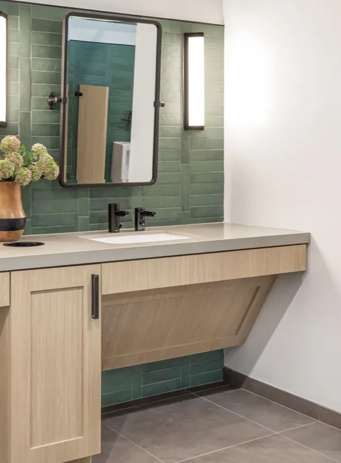 contemporary vanity with a light wood cabinet and beige countertop, featuring a black Sloan automatic faucet. A black-framed mirror is flanked by two vertical light fixtures, providing balance and symmetry. The back wall is clad in rectangular green tiles, adding texture and contrast.