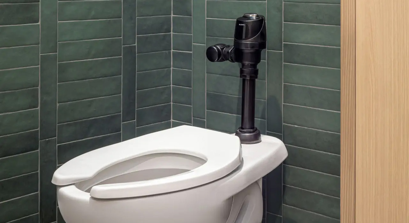 white porcelain toilet with a Sloan flushometer in a black finish, installed against a wall clad with vertically arranged green rectangular tiles. The contrast between the dark fixture and the light wood panel beside it creates an appealing visual balance.
