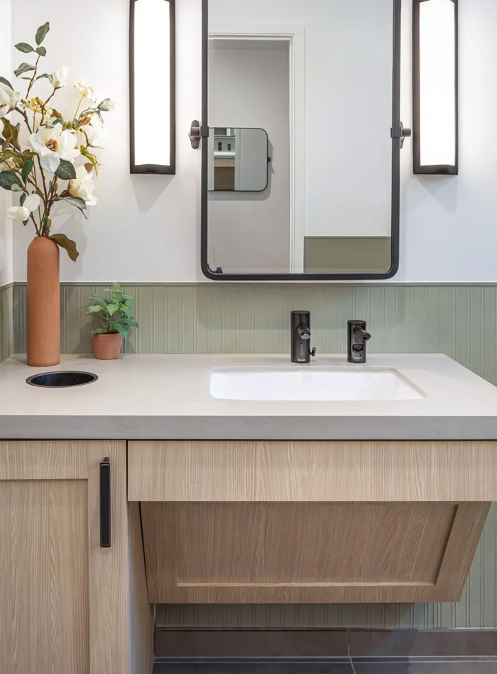 contemporary vanity with a light wood cabinet and beige countertop, featuring a black Sloan automatic faucet. A black-framed mirror is flanked by two vertical light fixtures, providing balance and symmetry. The back wall is clad in rectangular green tiles, adding texture and contrast.