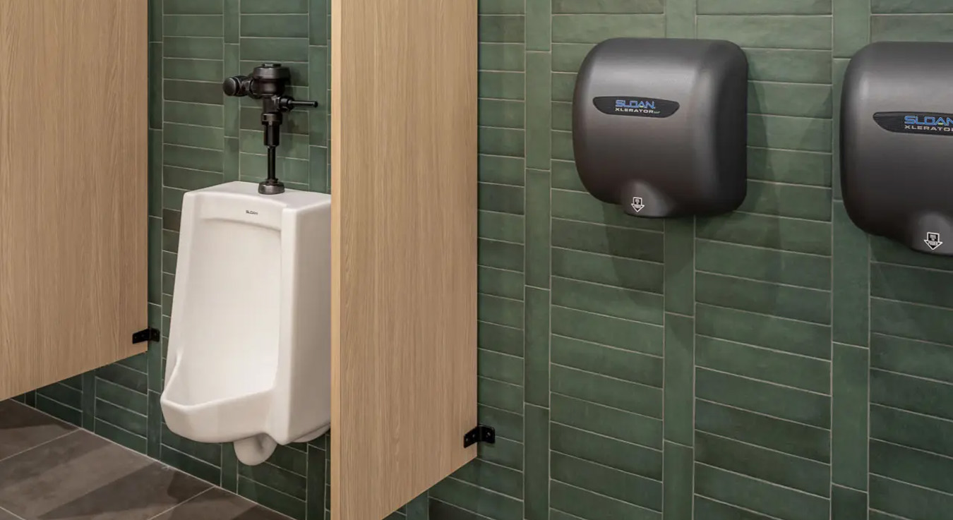 white urinal with a Sloan flushometer in a black finish, positioned between light wood privacy panels against a wall clad in elongated green tiles. To the right, Sloan automatic hand dryers in matching dark tones maintain visual consistency.