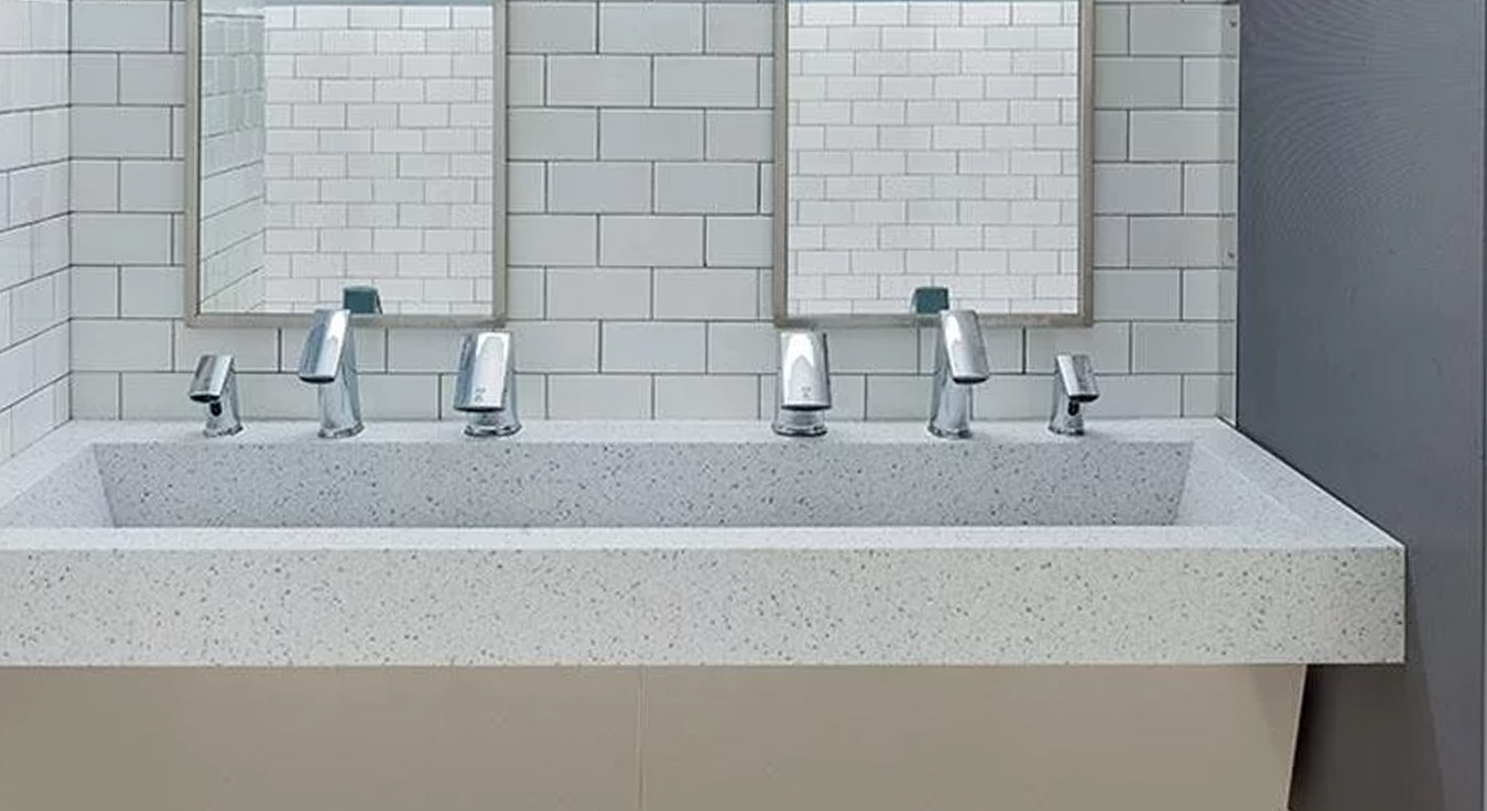 The image shows a contemporary restroom featuring multiple integrated solid-surface sinks equipped with Sloan automatic faucets. The white subway tile design and vertical mirrors create a clean, modern, and functional environment, ideal for high-traffic public spaces.