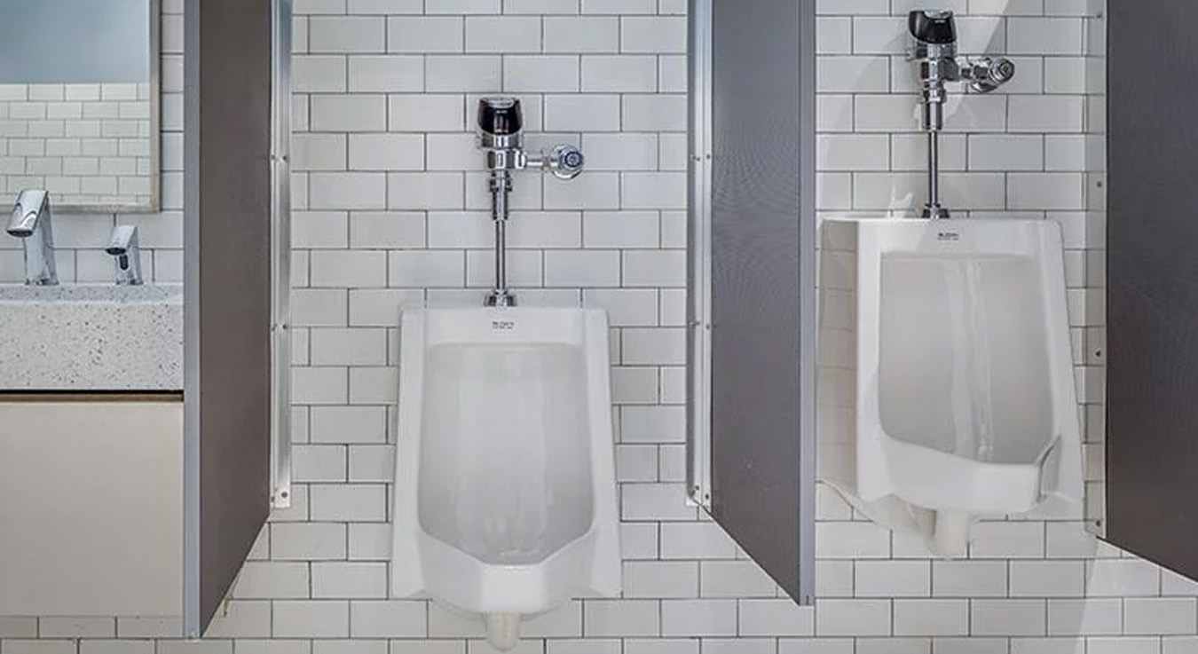 a modern public restroom featuring two Sloan urinals with chrome automatic flushometers and side partitions for added privacy. The design stands out with its white subway-tile walls and decorative flooring that adds a contemporary touch to the space.