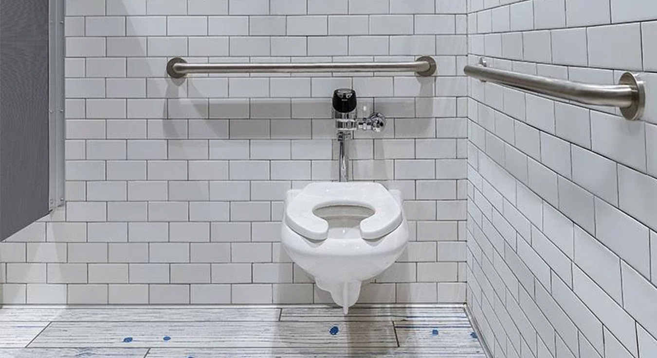 an accessible restroom designed with stainless steel grab bars on both sides of the toilet, meeting accessibility standards. The walls are covered with white subway tiles, and the floor features a decorative blue-toned design, adding a modern and clean touch to the space.