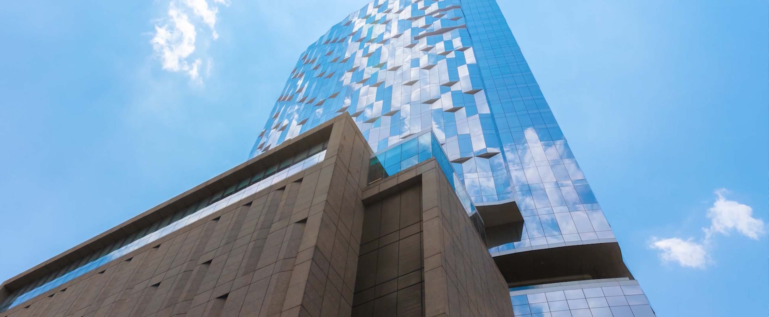 modern skyscraper with a reflective glass façade that mirrors the blue sky and clouds. At the base, brown-toned structures contrast with the metallic shine of the main tower, highlighting its contemporary and elegant design.