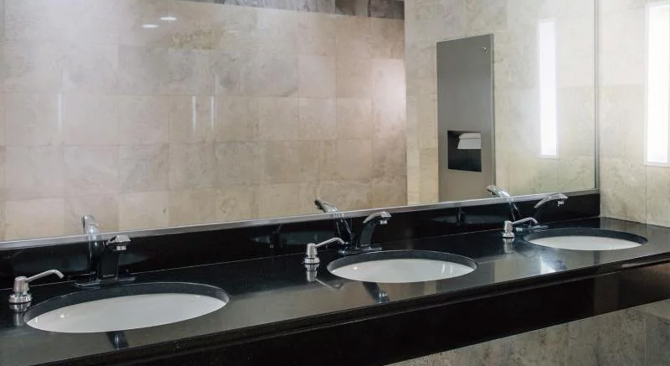 modern restroom featuring a black granite countertop with built-in sinks. A large mirror expands the space and reflects the recessed ceiling lights, creating a clean and elegant atmosphere. The light marble walls add a sophisticated touch to the overall design.