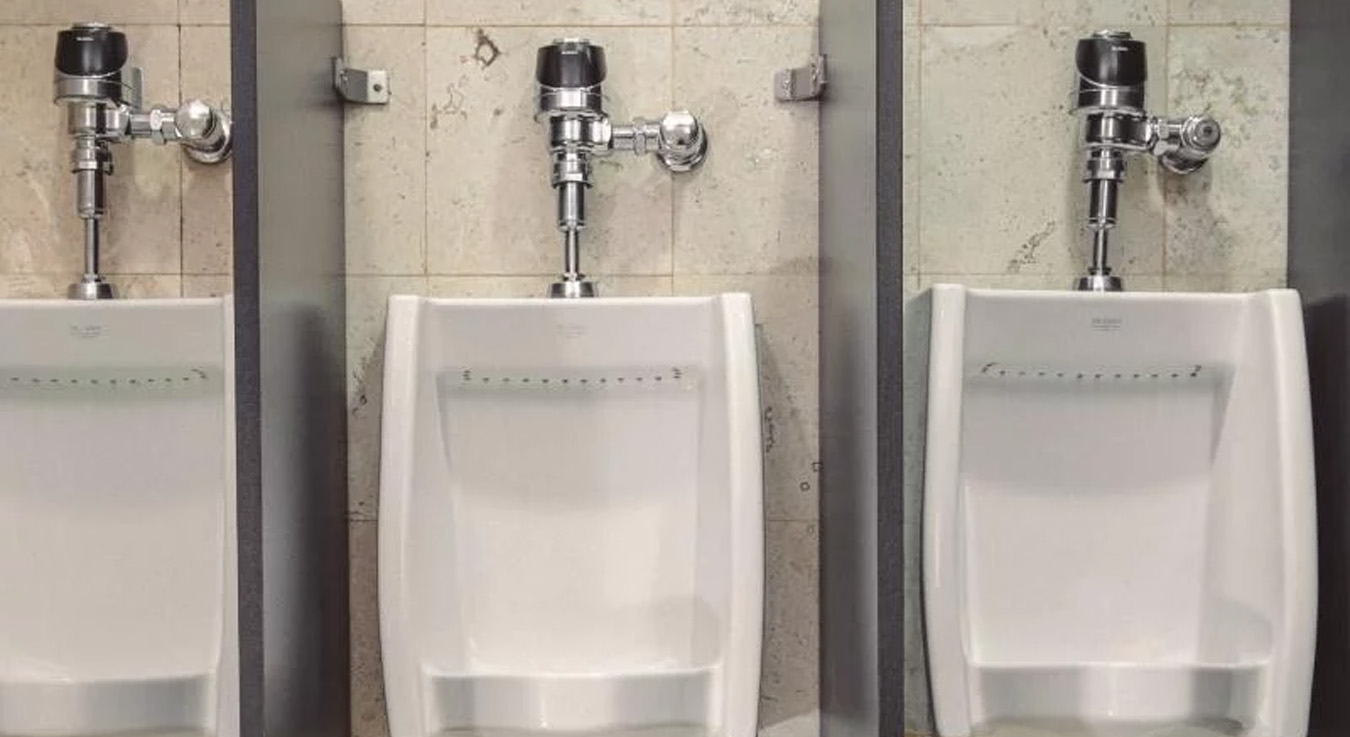 a row of three white urinals separated by gray panels, installed against a wall covered in beige and gray tiles. The design is clean and functional, featuring chrome flush valves that add a modern and durable touch.