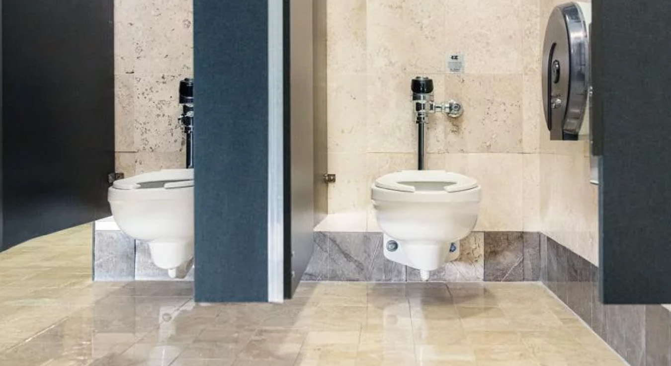 two restroom stalls with modern wall-mounted toilets, separated by gray panels. The walls and floor are covered with beige and marble-toned tiles, giving the space a clean and elegant appearance.