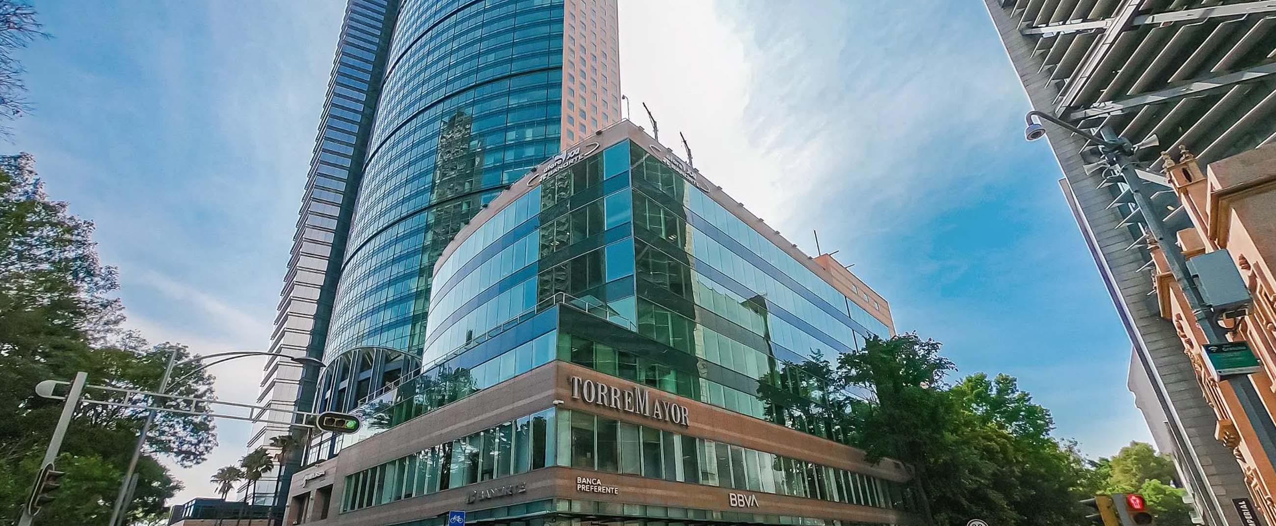 Torre Mayor, a modern skyscraper located in Mexico City. Its façade combines reflective glass and metal structures, standing out for its sleek design and impressive height. At the base, office spaces and bank branches are visible, while the surrounding urban environment conveys an orderly, contemporary atmosphere under a clear sky.