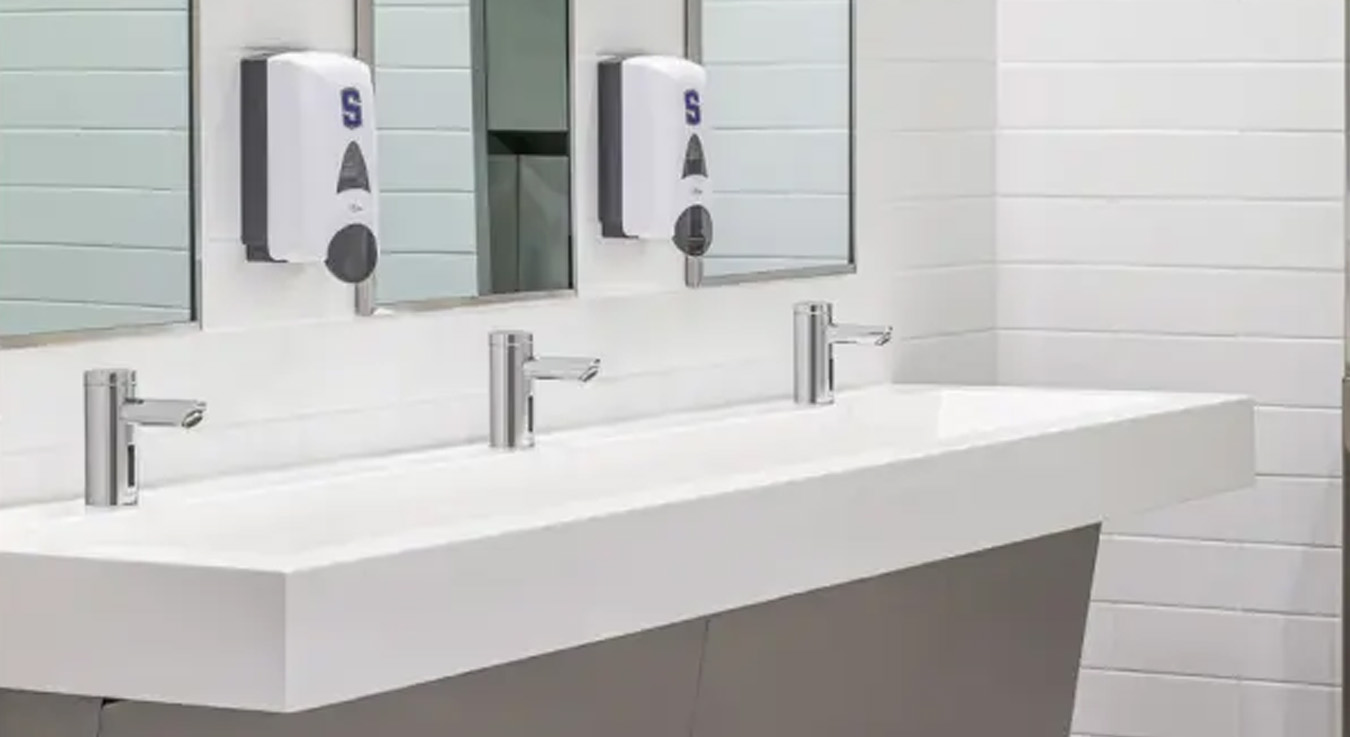 a multi-sink station equipped with Sloan automatic faucets, designed with a minimalist and functional aesthetic. The chrome finishes and solid white countertop convey cleanliness and modernity, while the touch-free soap dispensers enhance hygiene and user convenience
