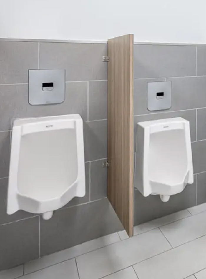 a set of wall-mounted Sloan urinals, each featuring a concealed electronic flush valve with a sleek stainless steel finish. This setup provides a clean, modern look with no exposed components, making it ideal for commercial and institutional facilities. A wooden privacy partition between the urinals adds warmth and discretion to the space. The neutral gray tile design enhances the professional, hygienic, and low-maintenance appearance of the restroom.