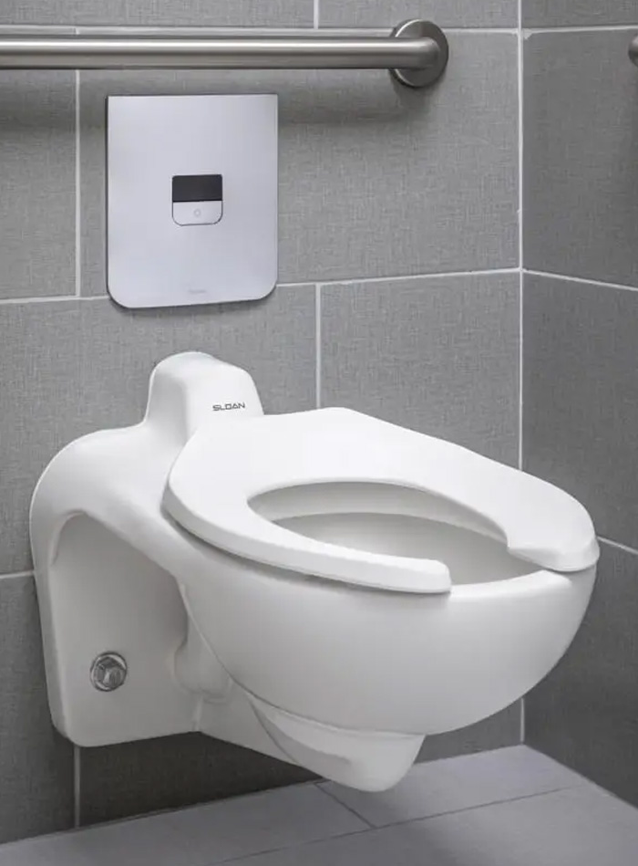 Sloan wall-mounted toilet installed in a modern commercial restroom. Its wall-hung design optimizes space and simplifies cleaning, while the concealed flush valve with stainless steel electronic panel delivers precise, touchless operation—perfect for high-traffic environments. The gray ceramic tile finish adds a clean, professional aesthetic, complemented by the stainless grab bar for accessibility and safety.