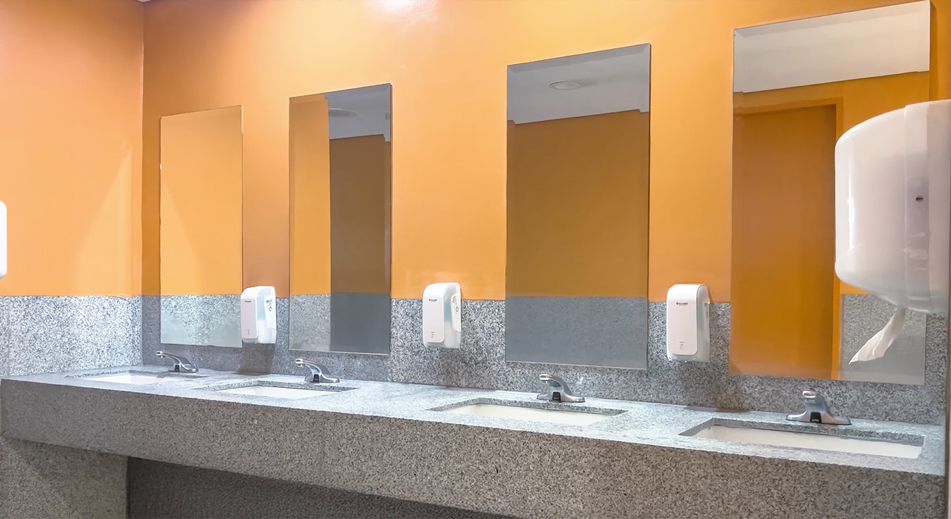 a public restroom with a clean and functional design. The walls are painted in a bright orange tone, while the sink area is covered with gray granite. Four sinks with chrome faucets, vertical mirrors, and automatic soap dispensers are visible, conveying a sense of order and cleanliness.
