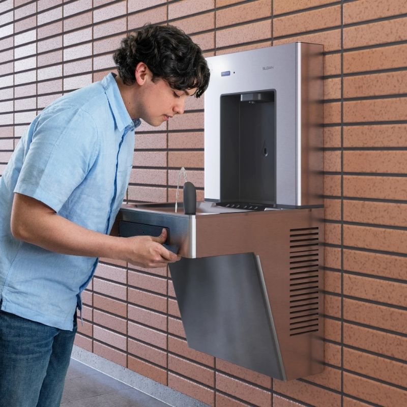 Person drinking from a wall-mounted Sloan drinking fountain with an integrated bottle filler, in a modern brick-walled setting.