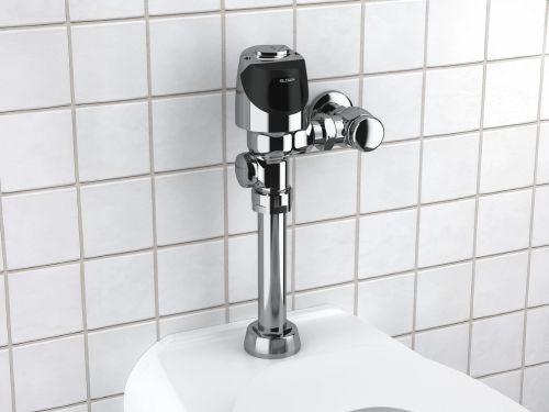 Válvula de descarga cromada instalada en un inodoro frente a una pared de azulejos blancos.